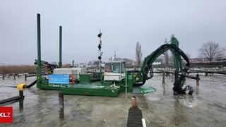 A large green industrial machine, labeled 'Classic V', is partially submerged in water near a dock, with a blue sign, traffic signals, and distant trees and buildings visible.
