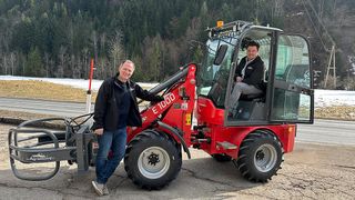 Zwei Männer in Winterkleidung stehen neben einem roten Traktor. Ein Mann steht, der andere sitzt im Fahrersitz des Traktors. Der Traktor ist auf einem ländlichen Gebiet mit verschneiten Boden und Bäumen im Hintergrund geparkt.