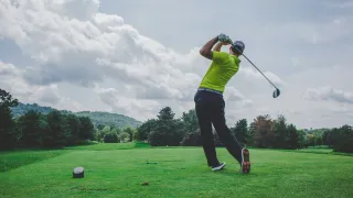 A golfer is swinging a golf club on a sunny day at a golf course. He is dressed in a green shirt, black pants, and white sneakers. There is a small golf tee on the grass.