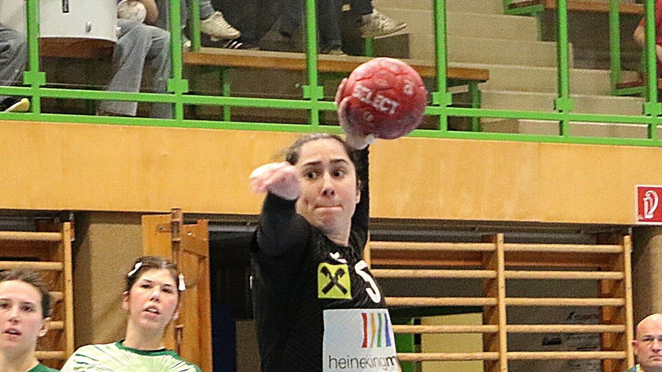 A woman in a jersey with the number 5 is throwing a red ball in a gymnasium. Spectators are seated on green benches above.