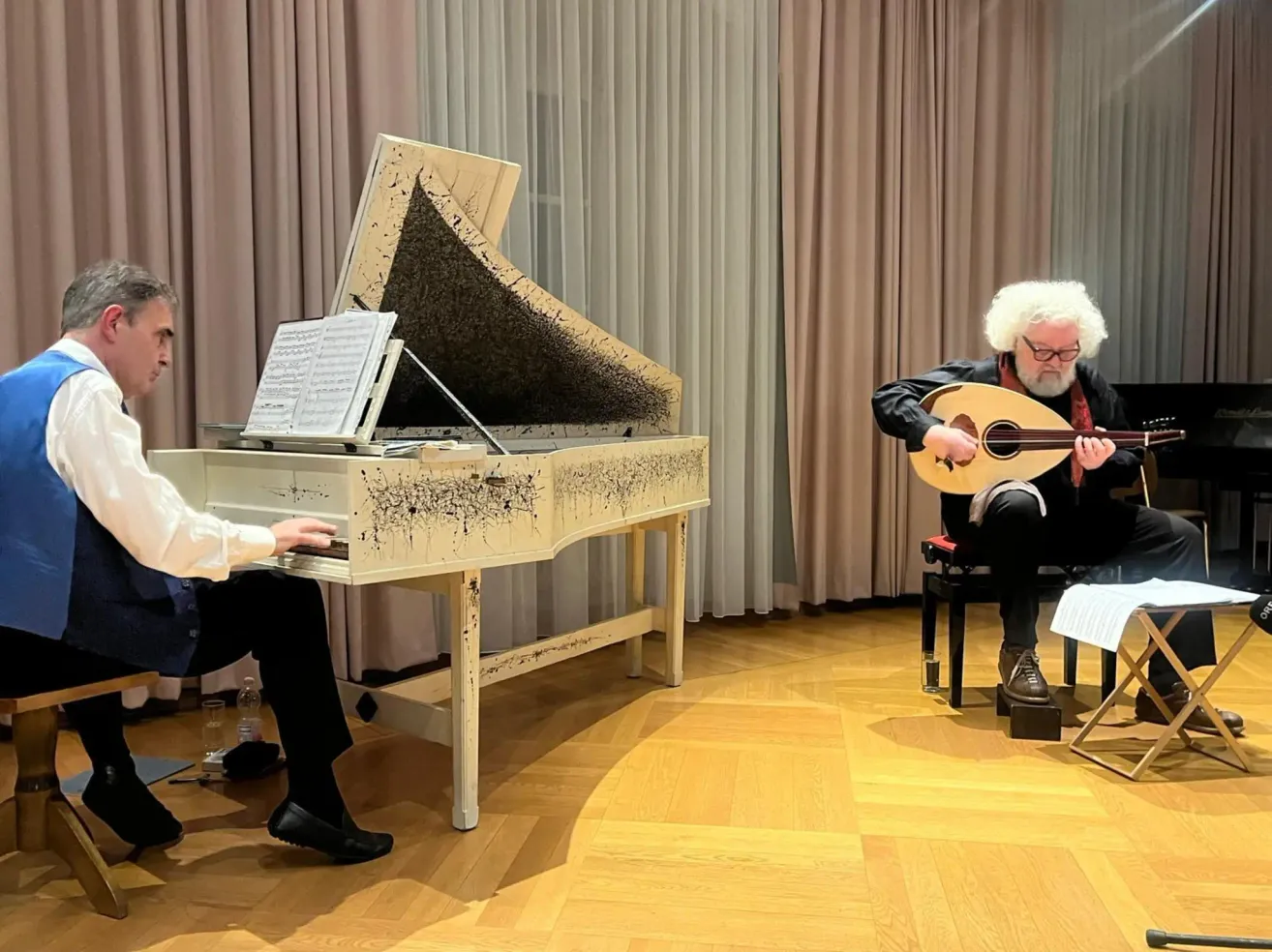 Two musicians are performing on stage. The man on the left is playing the piano, while the man on the right plays a lute.