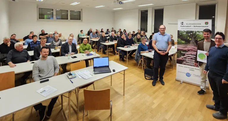 Ein Raum mit Holzboden und weißen Wänden enthält mehrere Personen, die an Tischen mit Laptops sitzen. Ein Mann steht vor ihnen, lächelt und trägt eine blaue Hemd.