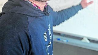 Eine Person in einer blauen Kapuzenjacke mit Design steht neben einer Tafel und zeigt mit einer Hand.