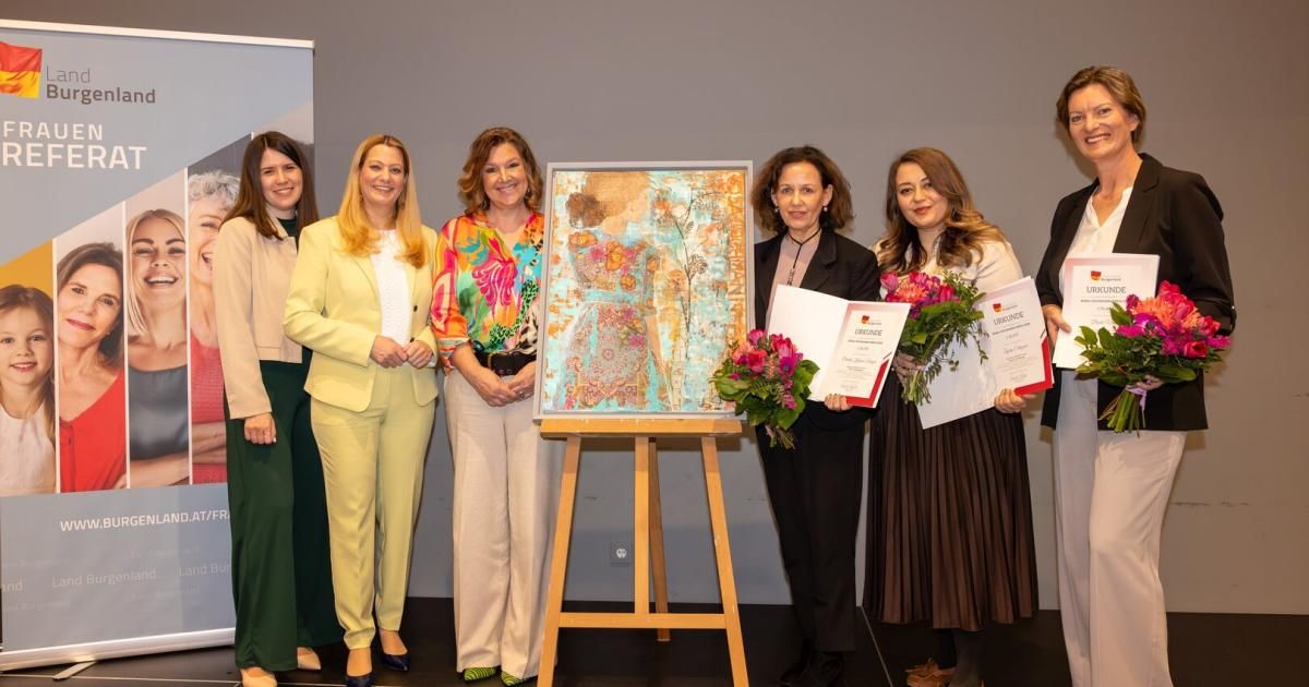Seven women stand in front of an art exhibit. Three hold certificates and flowers. The painting depicts a woman. Behind them is a banner.
