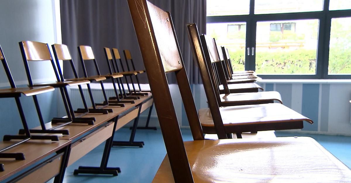 A row of brown wooden chairs are lined up in a classroom, facing away from the camera. The chairs have metal legs, and the floor is blue. A curtain and window are visible in the background.