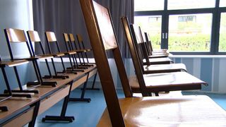 A row of brown wooden chairs are lined up in a classroom, facing away from the camera. The chairs have metal legs, and the floor is blue. A curtain and window are visible in the background.