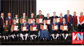 A group of people dressed in traditional attire, possibly for a celebration, sit on chairs on a stage holding certificates.