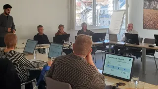 Fünf Personen sitzen an einem Tisch und schauen auf Bildschirme. Zwei Frauen und zwei Männer betrachten Bildschirme. Ein Mann mit kurzen blonden Haaren sitzt vorne. Ein Whiteboard ist im Hintergrund.