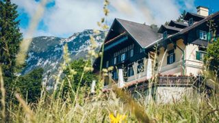 A building named Knappenhof stands in front of a mountain with a grassy field below. The building has a balcony with red flowers and a chimney on top.