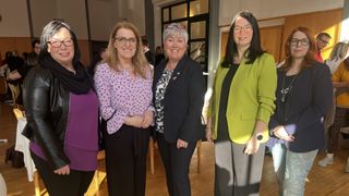 Four women are standing indoors, smiling for a photo. The woman on the left wears a purple blouse with black dots. The second from left has glasses and a black blazer. The third wears a green blazer and a watch. The fourth on the right wears a black top. Behind them is a wooden door.