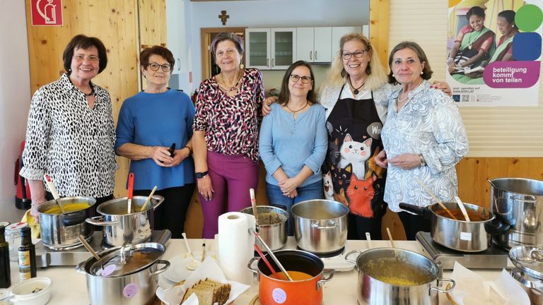 Five women in a kitchen, smiling, wearing aprons and necklaces, with pots and pans on the table. They stand behind a table filled with cooking utensils and ingredients.