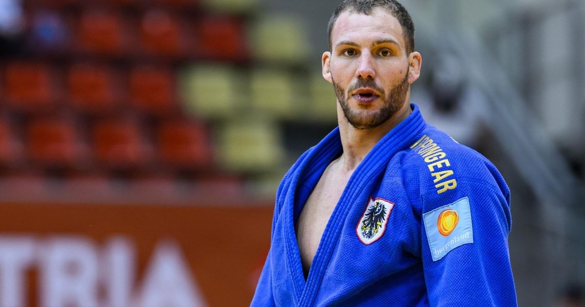 A man with a beard in a blue judo uniform, standing in a stadium, looking ahead.