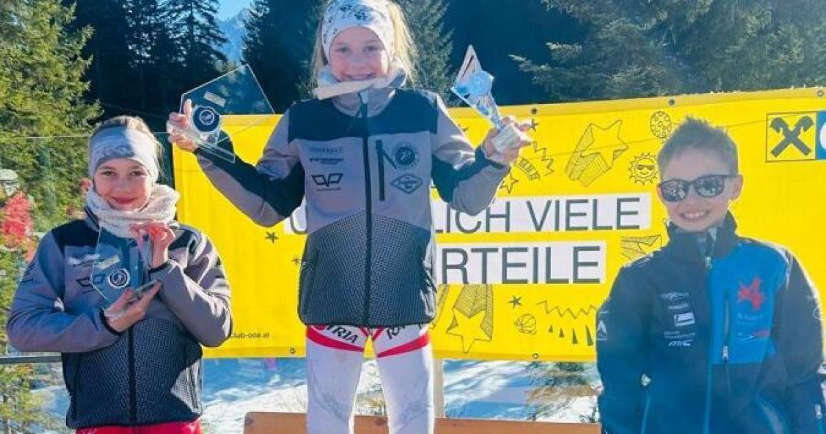 Ein junges Mädchen in einer Skiausrüstung steht auf einem verschneiten Podium und hält zwei Trophäen. Hinter ihr steht ein gelber Banner mit der Aufschrift 'ICH VIELE TEILE'. Zwei andere Kinder stehen neben ihr, eines trägt eine Brille. Bäume und ein verschneiter Hintergrund sind zu sehen.