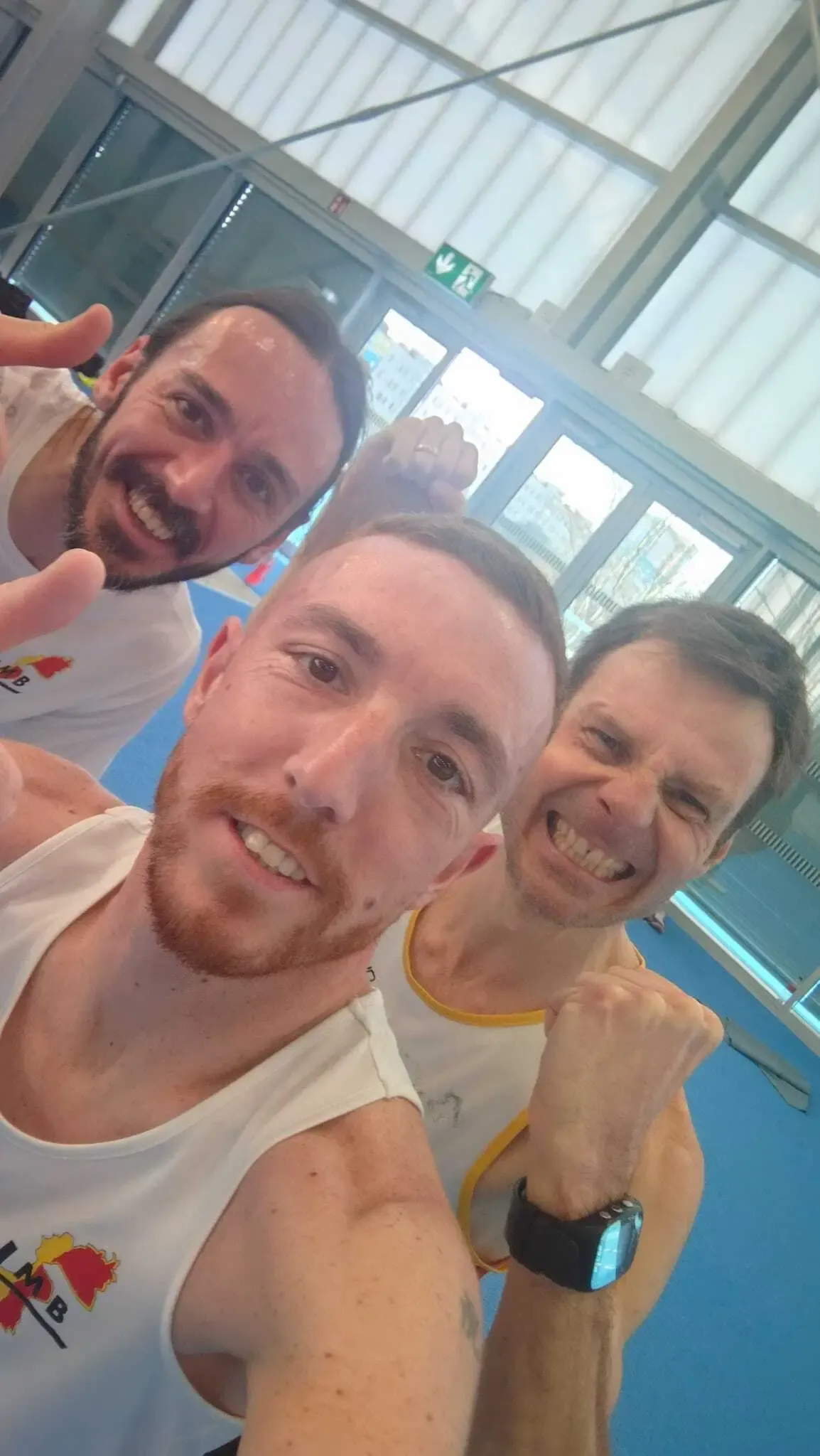 Three men are smiling and posing for a selfie in a gym. The man in the middle has a beard. The man on the right has a fist raised.