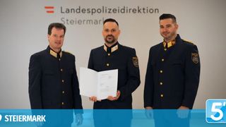 Three men in police uniforms stand in front of a white wall with a logo. The man in the middle holds a document, and the others stand beside him.