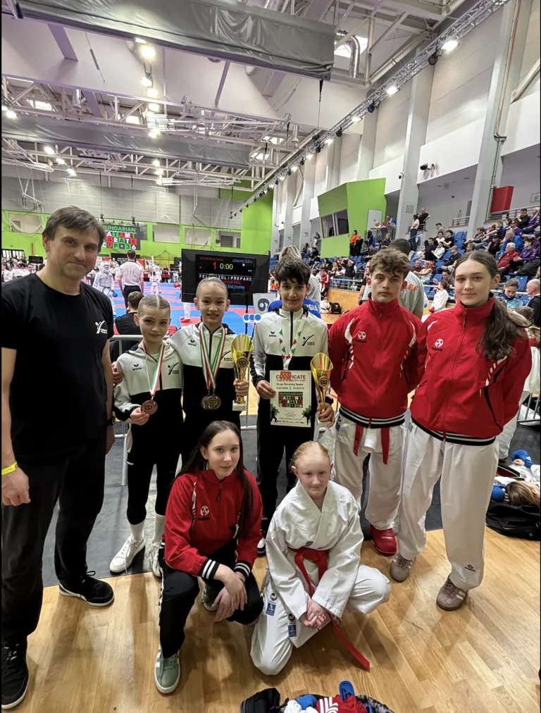 Five young athletes, three boys and two girls, are posing for a photo with medals and certificates. They are dressed in martial arts uniforms. Behind them, a scoreboard displays the number 1 and a time of 0:00. An audience is seated in the background.