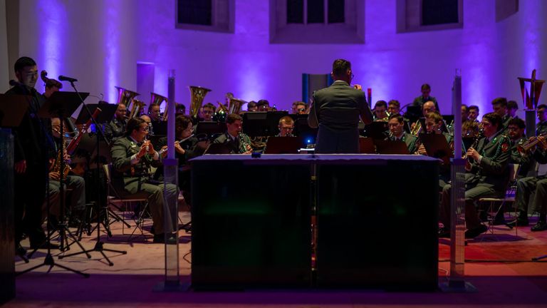 An orchestra is performing in a room with purple lighting. A man in a suit is standing in front of the musicians. The musicians are playing various instruments.