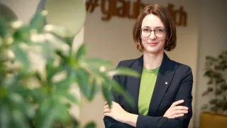 Eine Frau mit Brille und einem grünen Shirt posiert mit verschränkten Armen vor einer Wand mit einem verschwommenen Hashtag.