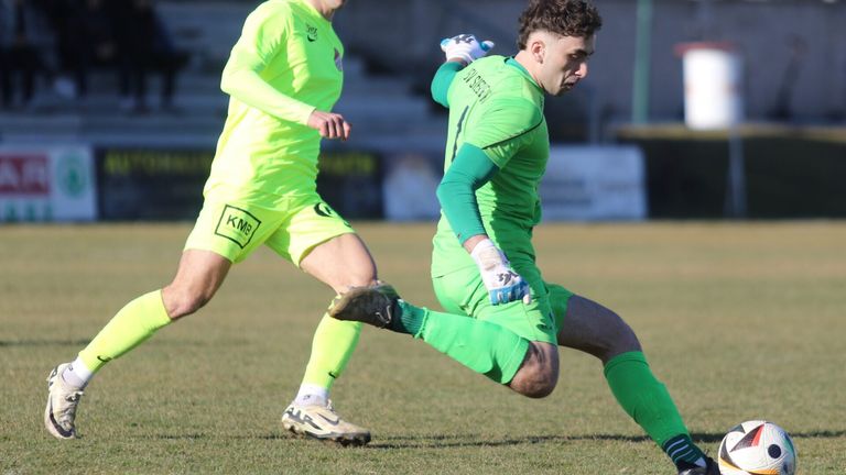 Two soccer players in green uniforms are on a field. One player is kneeling, and the other is running.