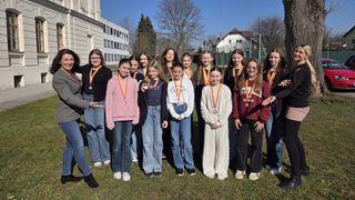A group of young girls wearing medals pose for a photo on a grassy field in front of a building.