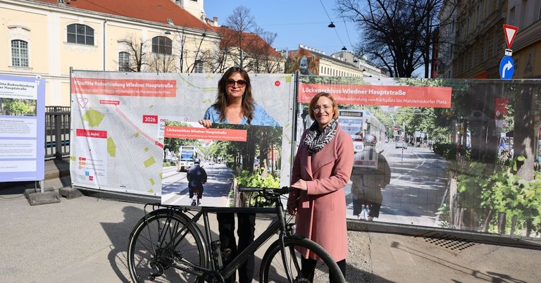 Zwei Frauen stehen neben einem Fahrrad mit einer großen Karte vor einem Banner, das eine Fahrradspur und ein Bild eines Straßenbahns zeigt.