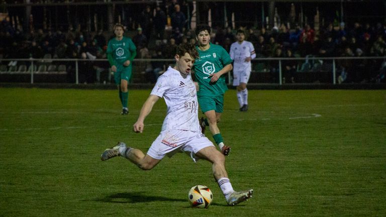 A soccer player in white kicks the ball as two players in green watch. The game is played at night on a field with spectators in the background.