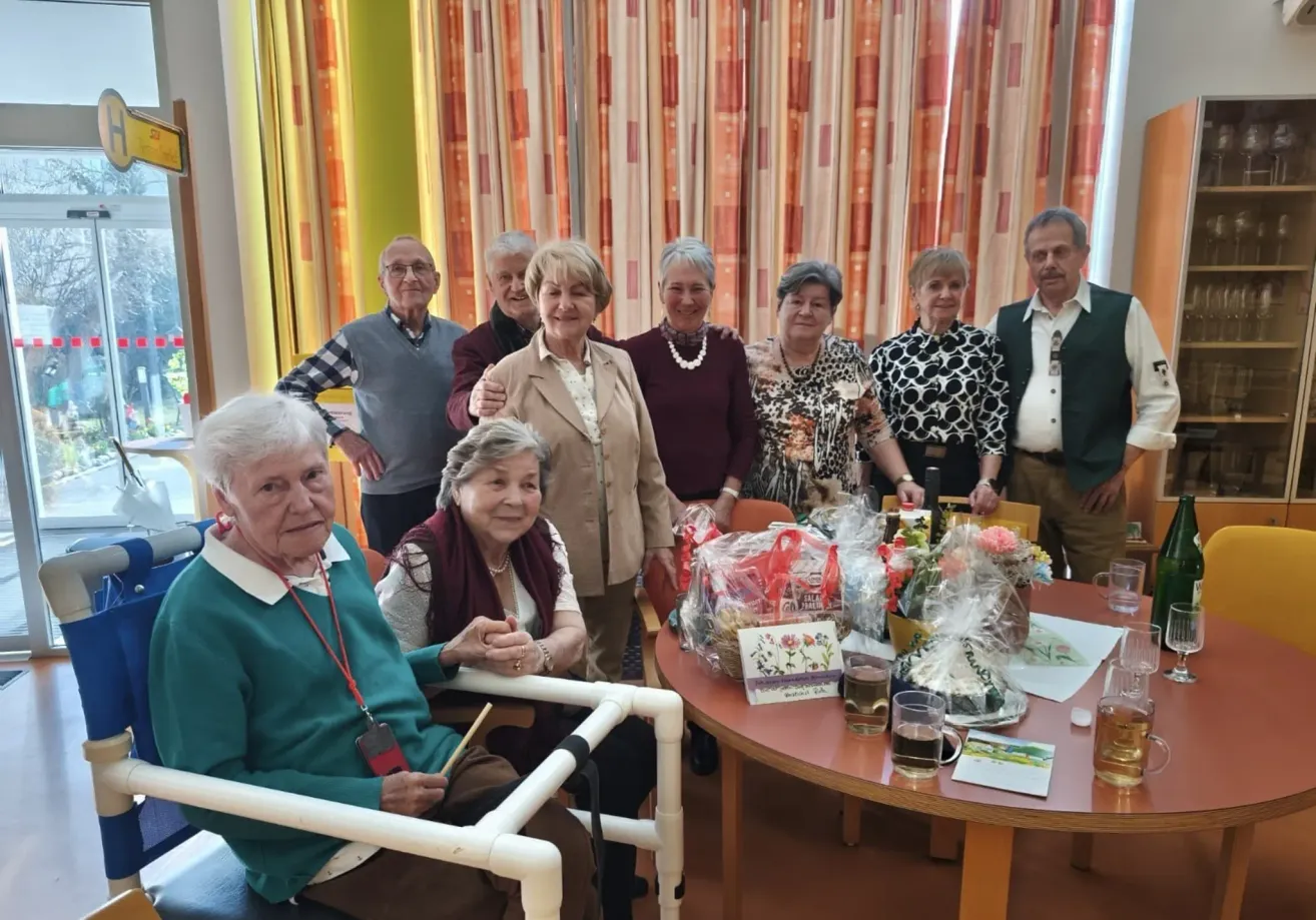 A group of elderly people are gathered around a table with gifts and flowers, smiling and posing for a photo in a room with curtains.