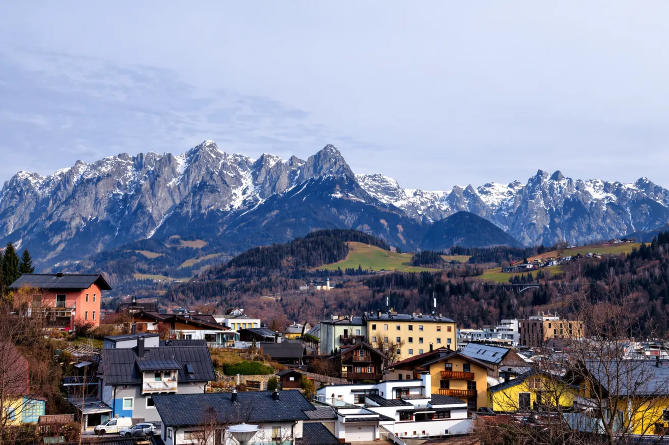 Eine Stadt mit schneebedeckten Bergen im Hintergrund, mit blauem Himmel und verstreuten Wolken. Die Stadt hat Häuser und Gebäude in verschiedenen Größen und Farben.