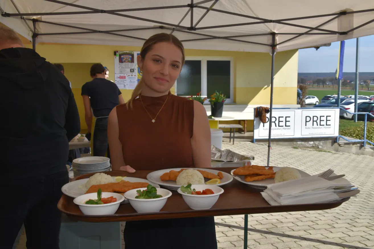 A woman stands under a tent, holding a tray of food. Several plates of food are on the tray, including fried fish and rice. A building and a car are visible in the background.