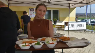 A woman stands under a tent, holding a tray of food. Several plates of food are on the tray, including fried fish and rice. A building and a car are visible in the background.