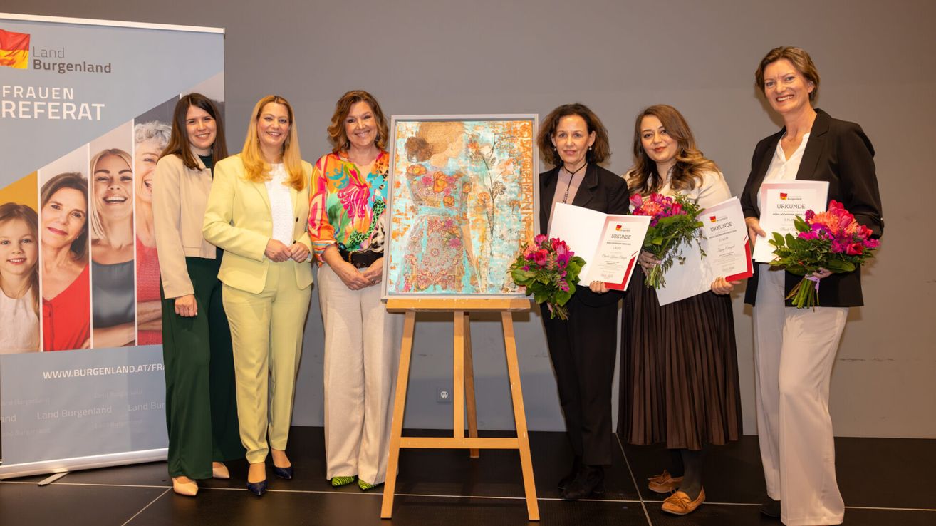 Five women stand on a stage holding certificates and flowers. A painting is displayed on an easel between them.
