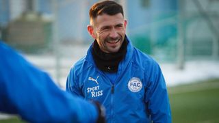 A man wearing a blue jacket with 'Sport Riiss' and a logo stands outdoors, smiling at someone off-camera.