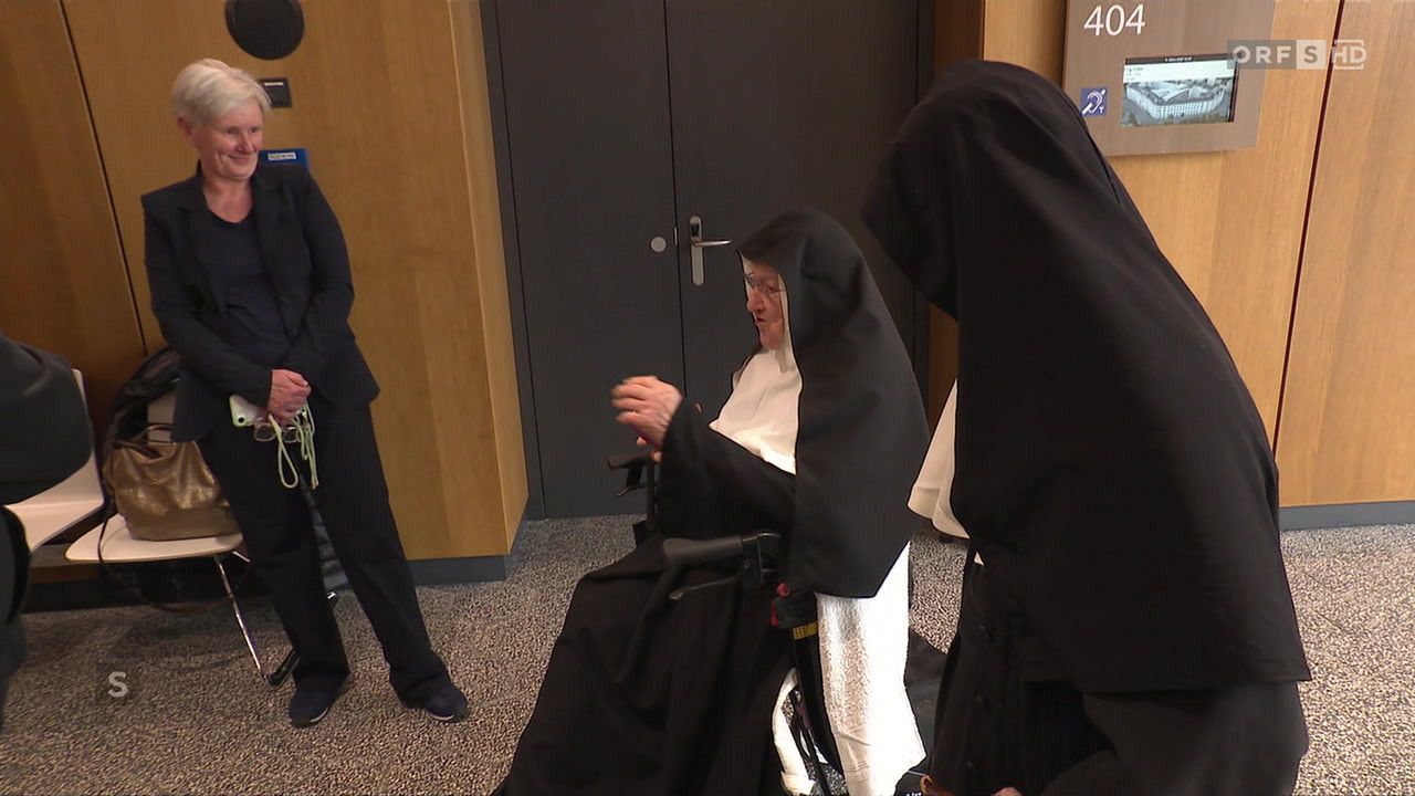 Eine ältere Nonne im Rollstuhl wird von einer anderen Nonne angeleitet, während eine Person in einem Anzug in der Nähe steht. Dahinter befindet sich eine Holzwand mit einer Tür.