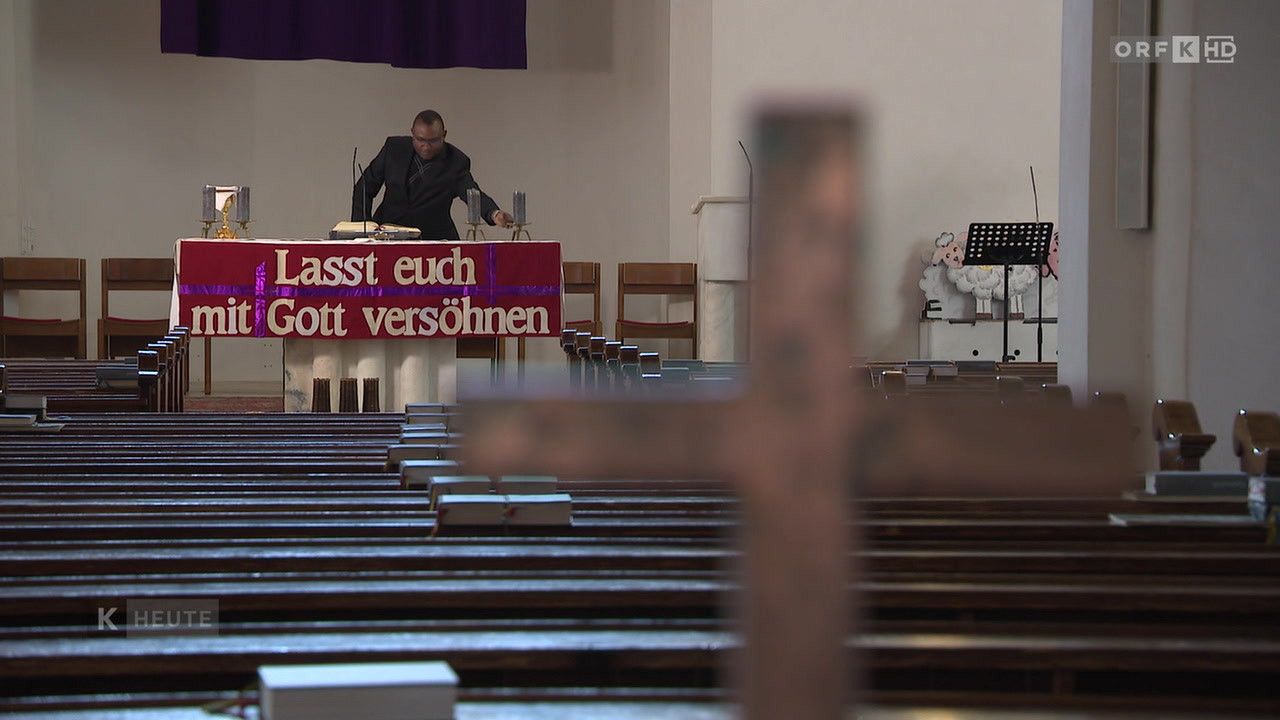 Eine Person steht an einer Kanzel in einer Kirche. Ein rotes Banner trägt die Aufschrift 'Lasst euch Gott versöhnen'. Die Kirchenbänke sind leer.