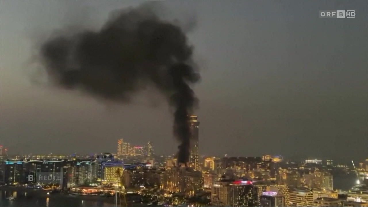 A city skyline at night with a large building on fire, emitting thick black smoke. The city is illuminated by numerous lights.