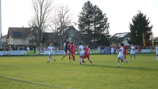 A soccer game is in progress on a field with players from two teams. One player jumps to head the ball. The field is surrounded by houses and a crowd of spectators.