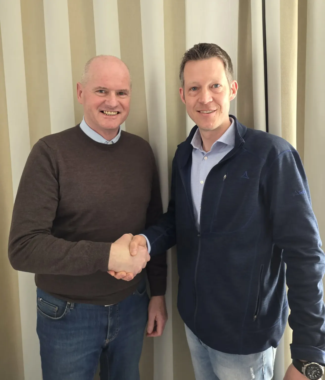 Two men are standing next to each other and shaking hands. They are smiling and appear to be posing for a photo.