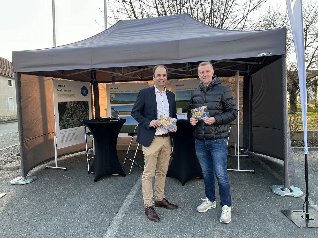 Zwei Männer stehen unter einem Zelt und lächeln für die Kamera. Einer hält ein Buch und der andere eine Broschüre. Sie befinden sich in einem Parkplatz mit einem schwarzen Baldachin.