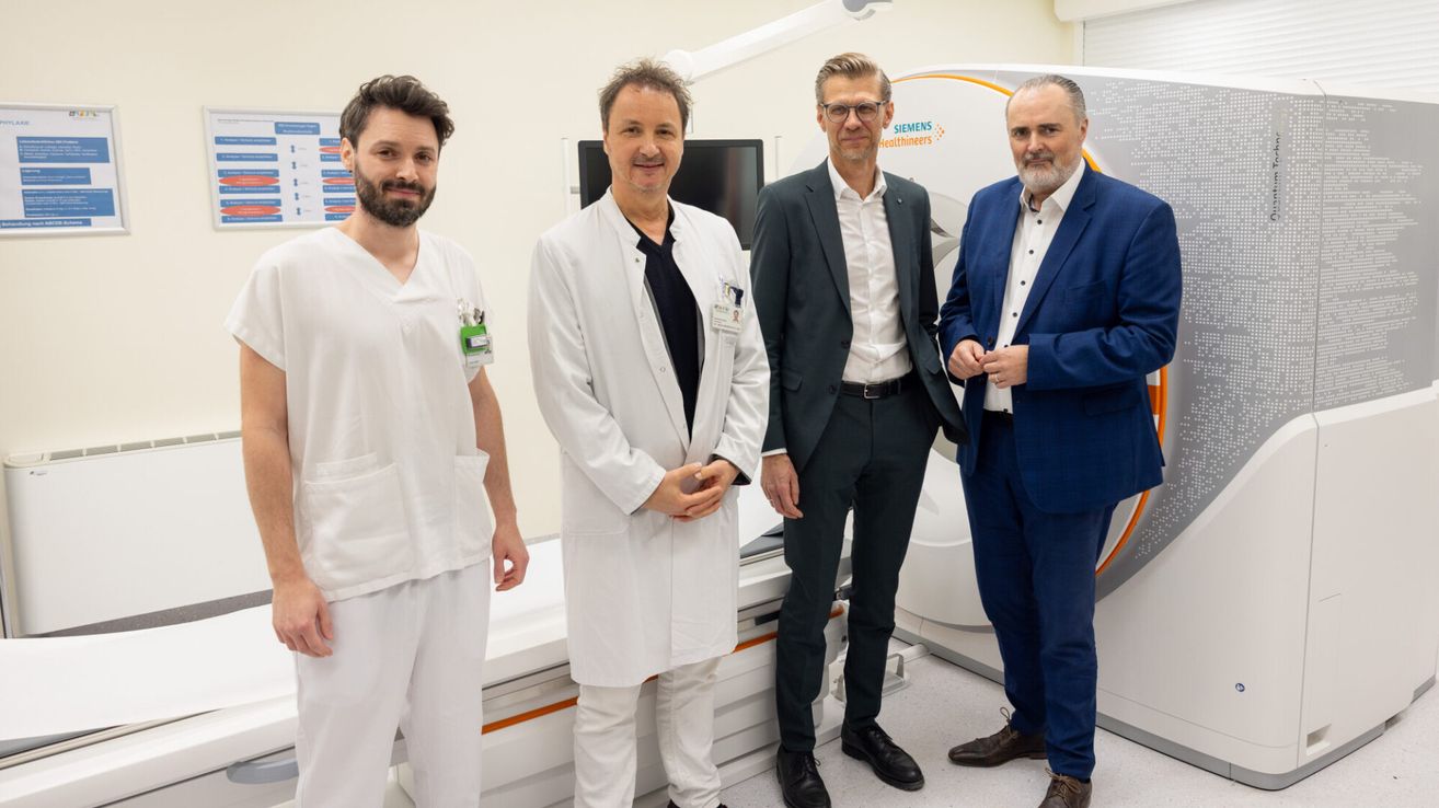 Four men stand in a hospital room near a Siemens Healthineers machine. Three wear suits, and one wears a white lab coat.