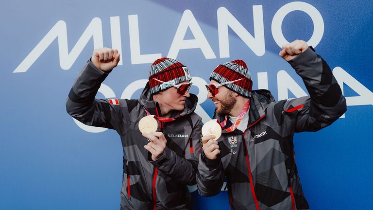 Two men wearing winter jackets, goggles, and hats celebrate with gold medals in front of a blue wall with text.