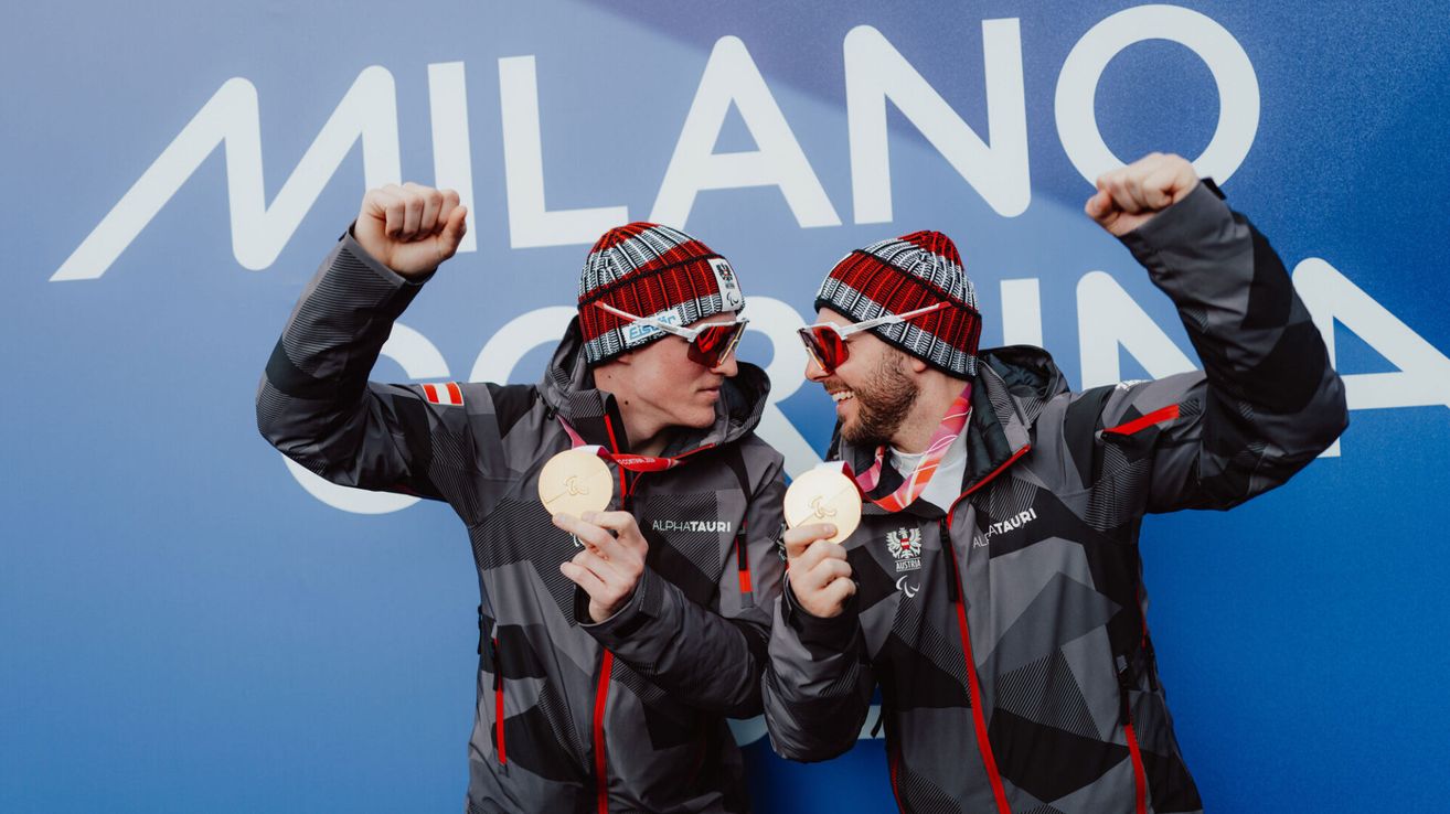 Two men wearing winter jackets, goggles, and hats celebrate with gold medals in front of a blue wall with text.