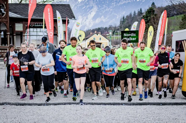 A group of runners wearing green and orange shirts and numbered bibs are participating in a marathon in a scenic area with mountains in the background.