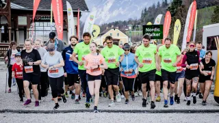 A group of runners wearing green and orange shirts and numbered bibs are participating in a marathon in a scenic area with mountains in the background.