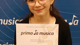 A young girl with glasses smiles while holding a certificate that reads 'prima Ja musica'. The background is blue with musical notes.