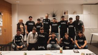 A group of people wearing black t-shirts with the word SCRA and smiling. A woman in fur and a man in a white shirt are also present. They are in a room with a table, pencils, and a plant.