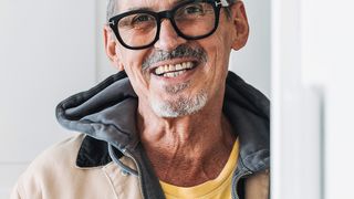 A man wearing a yellow shirt, a beige jacket, and black glasses stands against a white wall, smiling.