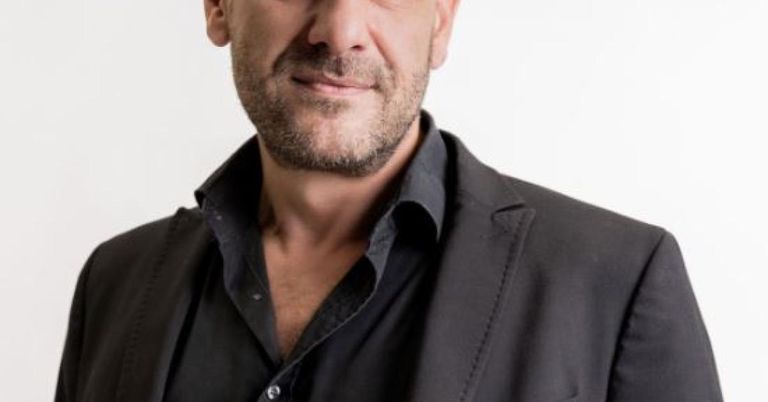 Ein Mann mit Bart und Schnurrbart lächelt, trägt ein schwarzes Hemd und eine Jacke und steht vor weißem Hintergrund.