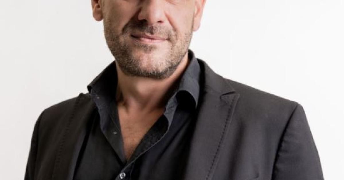 Ein Mann mit Bart und Schnurrbart lächelt, trägt ein schwarzes Hemd und eine Jacke und steht vor weißem Hintergrund.