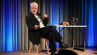 An older man with a white beard sits on a stool, holding a glass of water, and appears to be engaged in conversation. Behind him, there is a table with a vase and a cloth.