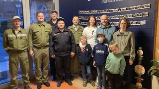 A group of individuals in uniforms and casual clothes pose for a photo in a room with a blue wall adorned with pictures.